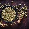 Whole cardamom pods overflowing a black bowl with a wooden spice scoop on a dark table. 