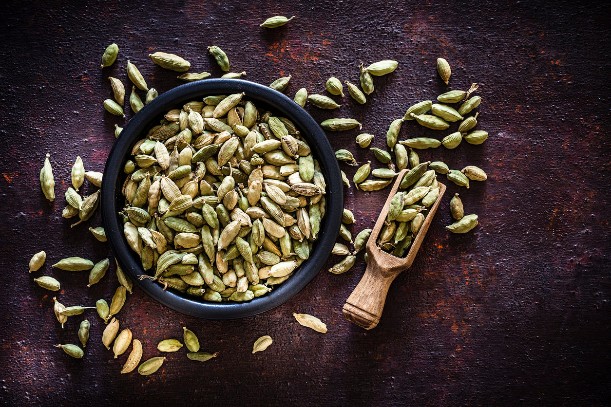 Whole cardamom pods overflowing a black bowl with a wooden spice scoop on a dark table. 
